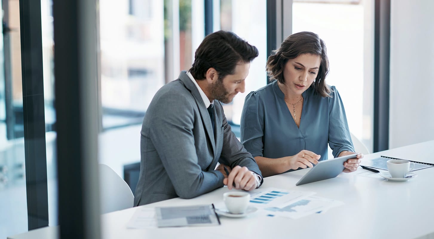 Two business professionals seated at a table in an office, collaborating over a tablet device.